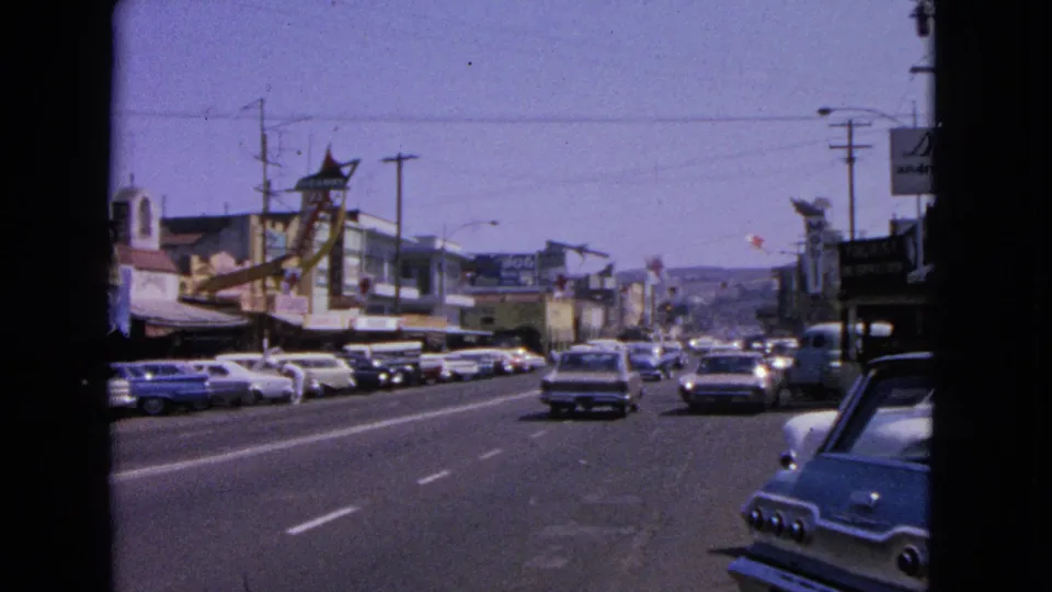Early-1960s traffic in Barrington, Illinois, with a car receding down the road, parked vehicles, storefronts, and visible 8mm frame borders.