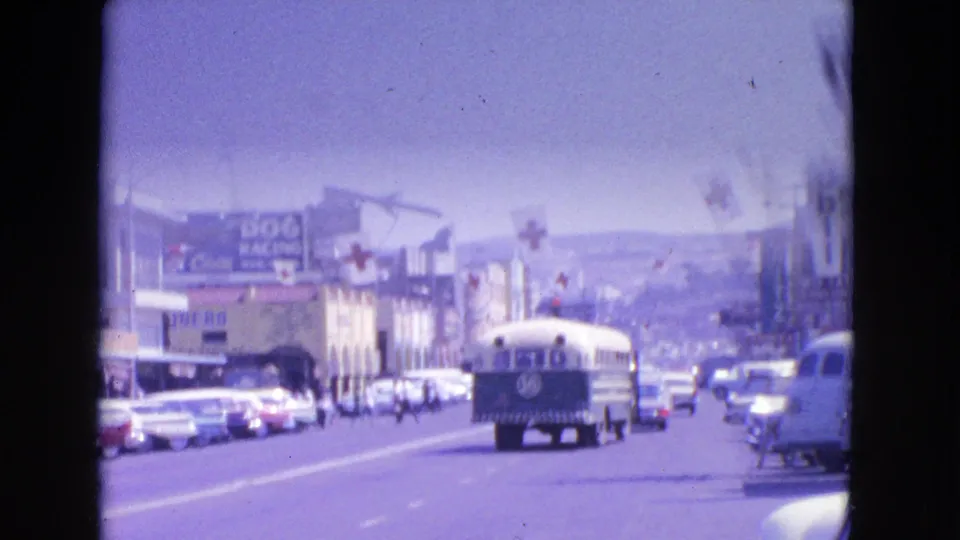A school bus travels along a 1963 Barrington, Illinois commercial street, with cars, storefronts, large signs, and faded 8mm color visible.