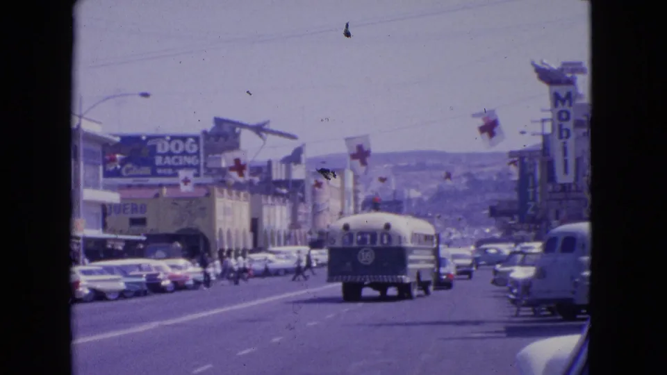 1963 Barrington, Illinois street view with a bus marked 16, classic cars, overhead cross flags, storefronts, and visible Mobil and dog racing signage.