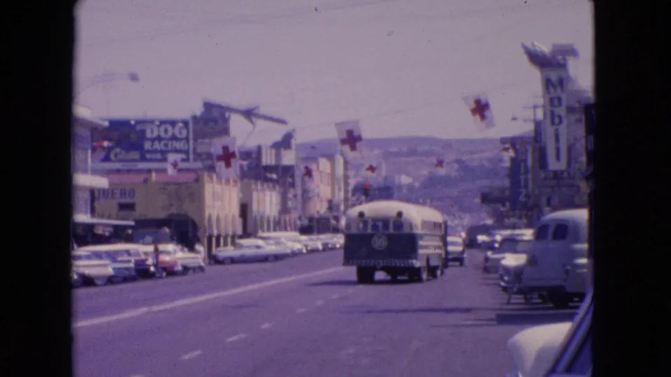 1963 Barrington, Illinois traffic view with classic cars parked and moving, a bus ahead, pedestrians near the curb, and cross flags above the road.