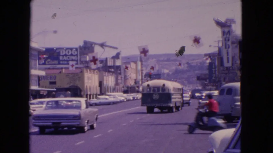 1963 Barrington, Illinois street frame with a motorcycle crossing the foreground, a bus ahead, classic cars, overhead cross flags, and visible roadside signs.