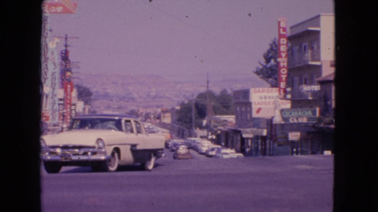 A 1963 8mm view in Barrington, Illinois, showing a white sedan turning past storefronts, hotel signage, and distant traffic.