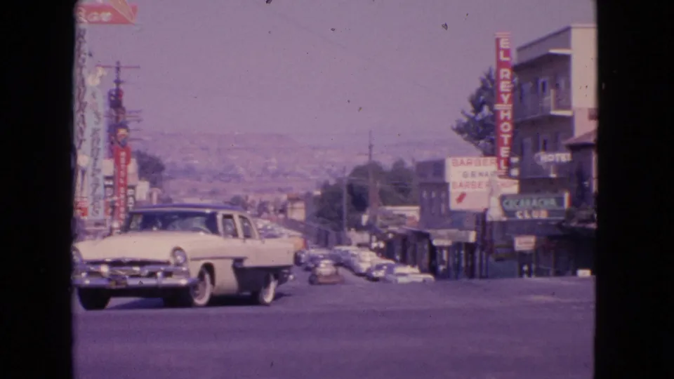 A 1963 8mm view in Barrington, Illinois, showing a white sedan turning past storefronts, hotel signage, and distant traffic.