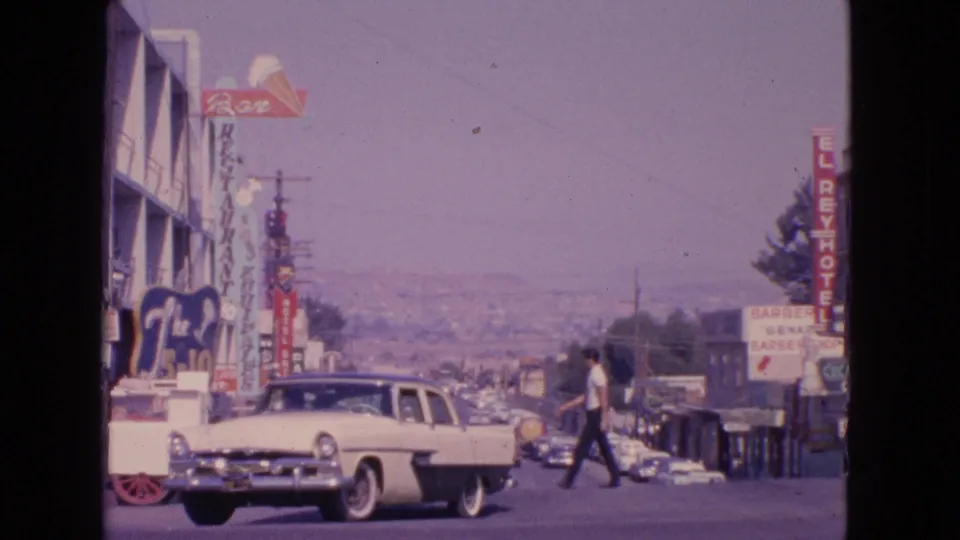 A pedestrian crosses in front of a white sedan in this 1963 Barrington street view, with hotel and shop signs behind him.