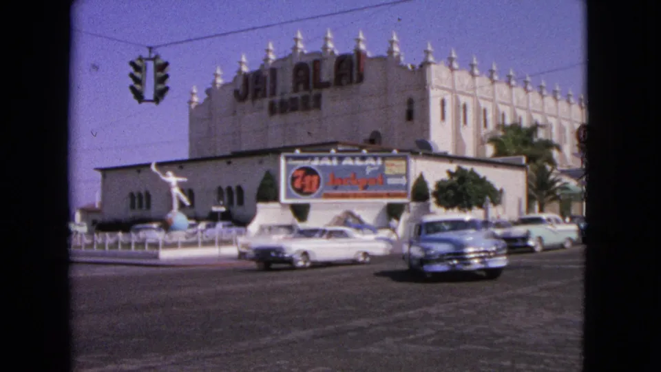 1963 8mm frame of classic cars crossing the foreground near prominent Jai Alai signage and a decorative facade.