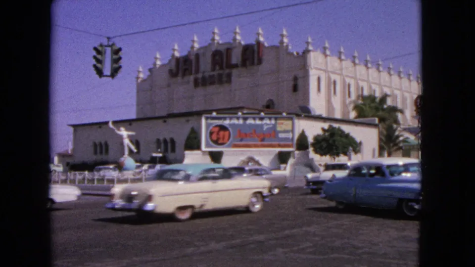 1963 home-movie frame with cream and blue cars moving through an intersection beside a Jai Alai building exterior.