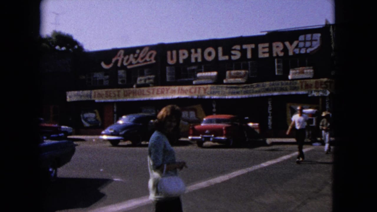 Pedestrians and parked classic cars outside the Avila Upholstery storefront.