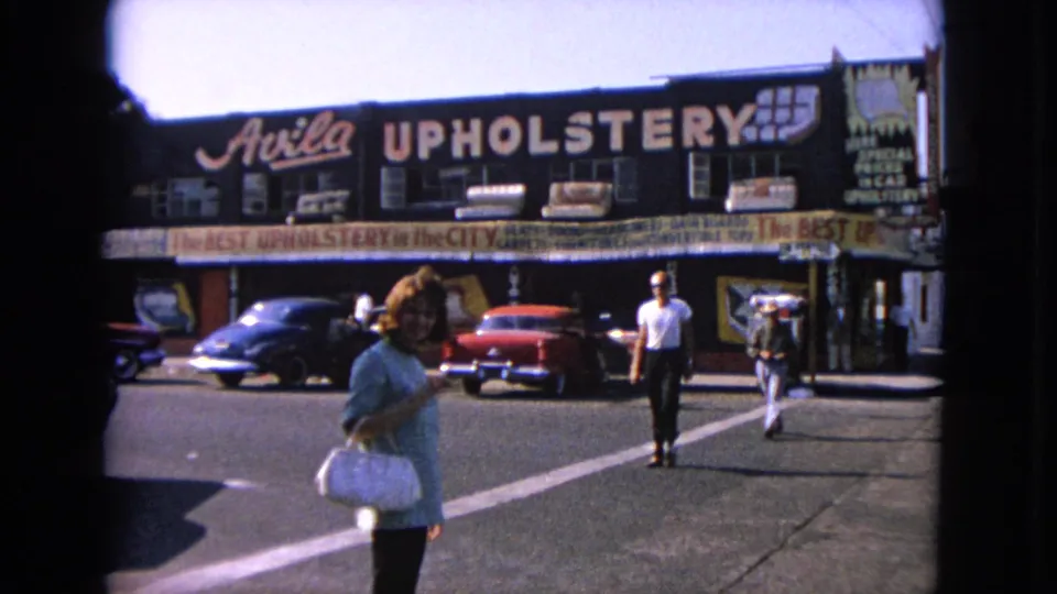 A woman with a white handbag stands near the camera while pedestrians cross in front of Avila Upholstery and classic cars.