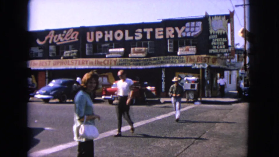 1963 Barrington street corner with pedestrians crossing, Avila Upholstery signage, parked classic cars, and strong midday shadows.