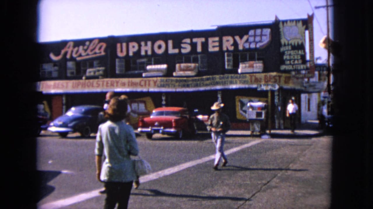 Pedestrians crossing in front of an Avila Upholstery storefront with classic cars parked nearby.