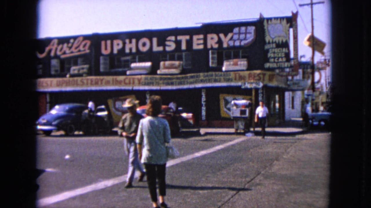 Two pedestrians cross the street toward a large upholstery storefront sign.