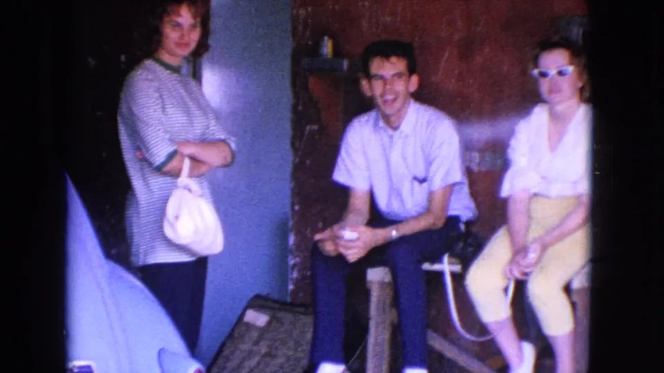 A brief indoor home-movie frame shows three adults near a wall or booth, including one standing woman and two seated people facing the camera.
