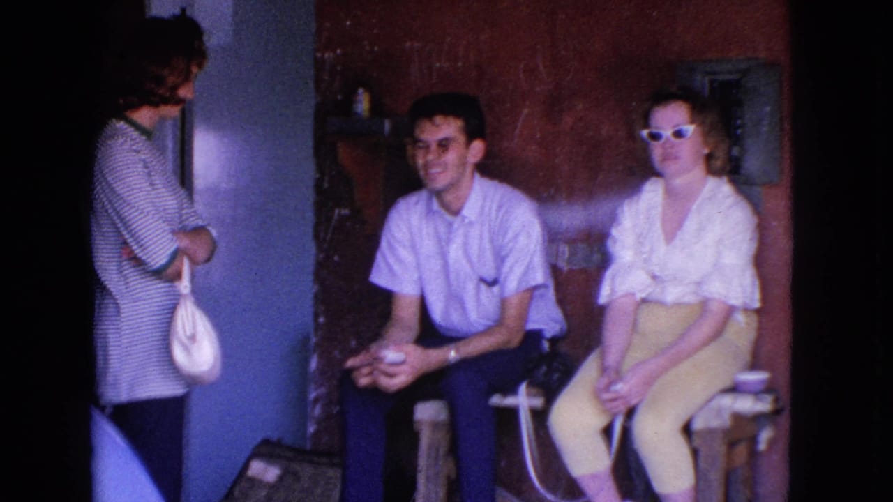 Barrington, Illinois 1963 home-movie frame showing two seated adults and a standing adult in a dim reddish-walled interior.