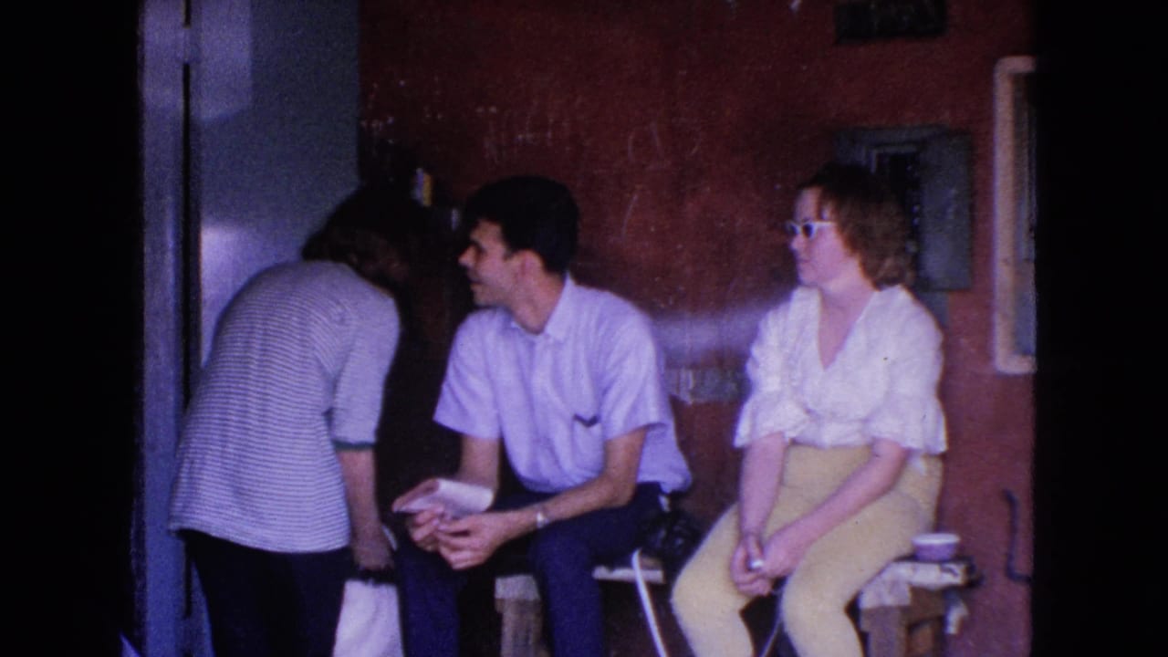 1963 Barrington home-movie frame with a standing adult leaning toward a seated man and woman in a dim interior room.