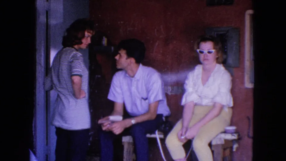 1963 Barrington 8mm frame of three adults gathered near a bench and doorway in a dim private interior.