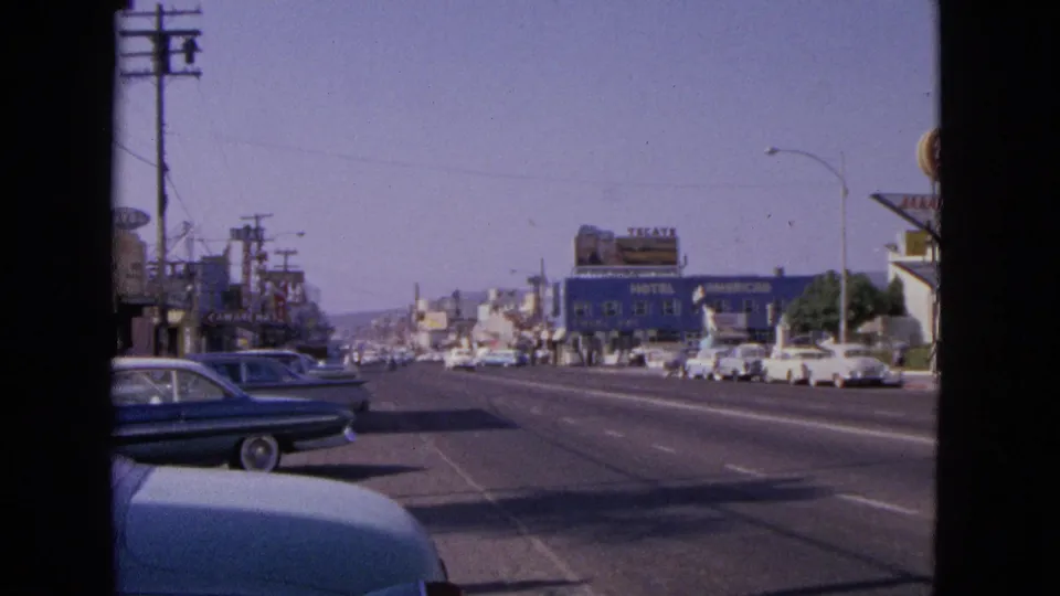 1963 Barrington roadside view with a blue sedan in the foreground, utility poles, motel signage, and commercial buildings beyond.