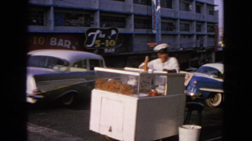 1963 Barrington, Illinois sidewalk scene with a man in a white cap behind a food cart, classic cars, and The 5-10 Bar sign.