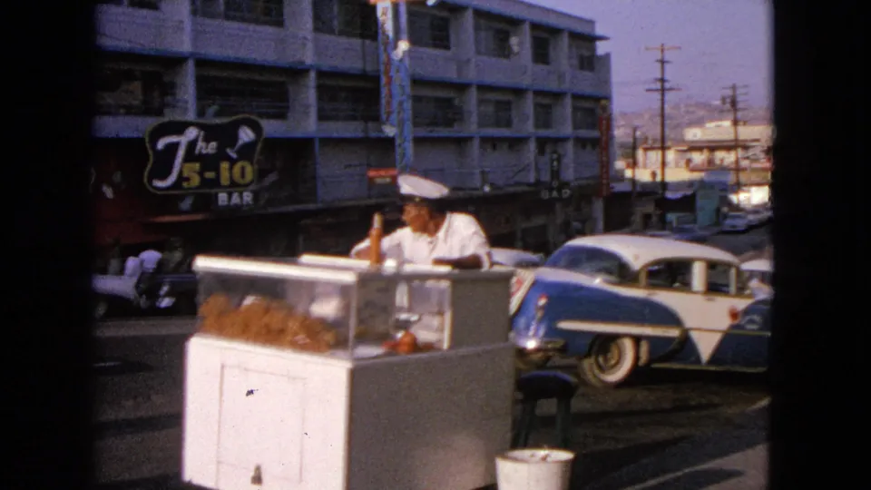 1963 Barrington, Illinois street vendor beside classic cars and storefront signage, with utility poles and distant terrain beyond the road.