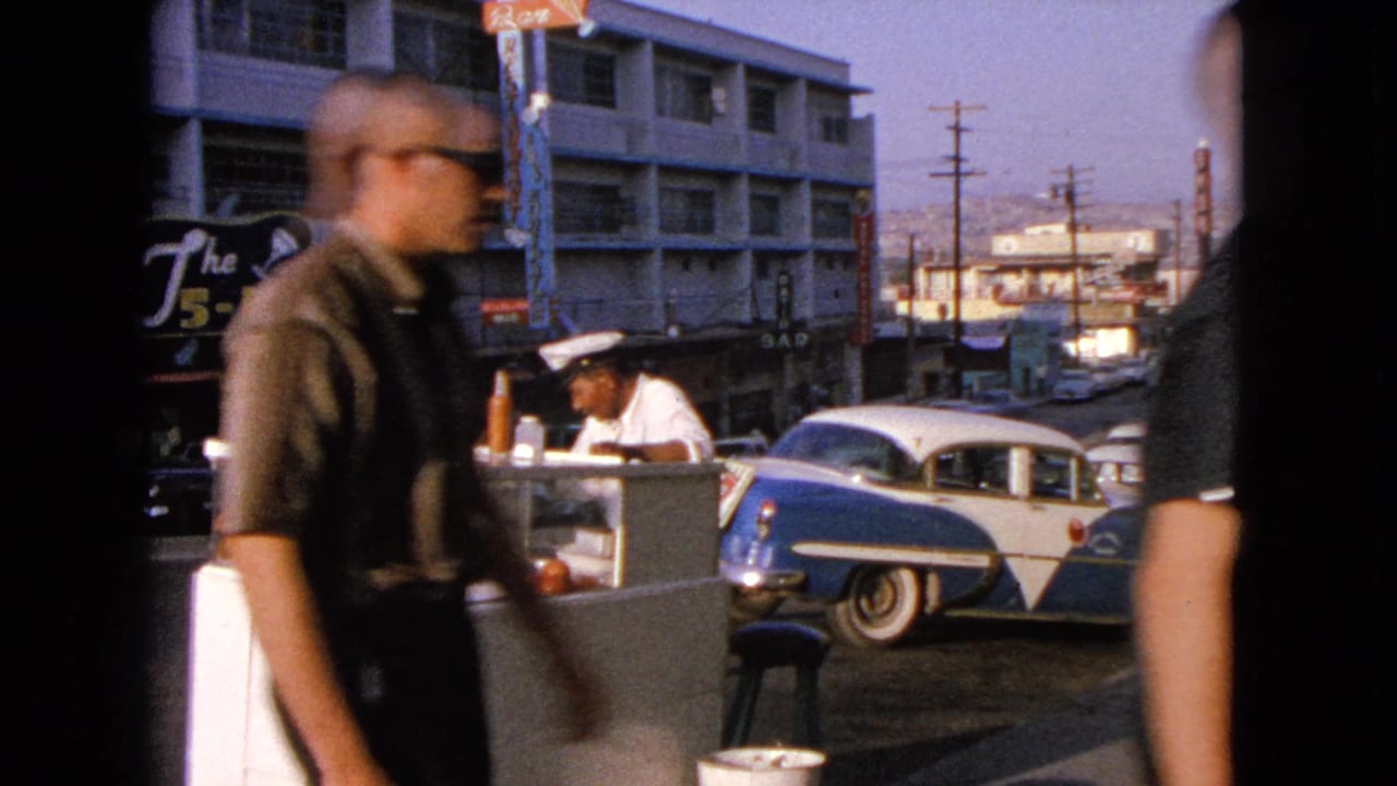 Pedestrian in sunglasses crosses the 1963 Barrington sidewalk scene, partly obscuring the vendor, hot dog stand, and blue-and-white sedan.