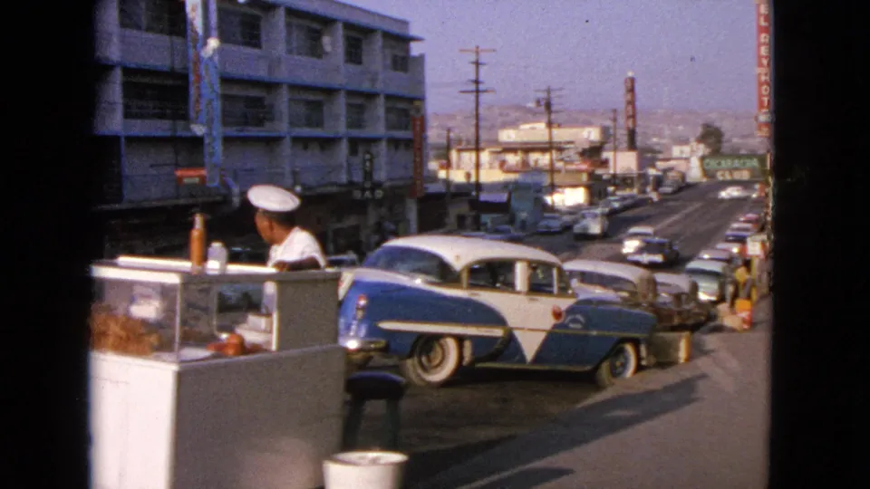 Wider 1963 Barrington view showing the hot dog stand, vendor, parked blue-and-white sedan, moving traffic, storefronts, and El Rey Hotel sign.