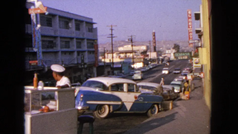 1963 Barrington street frame with a white hot dog stand in the foreground, vendor in cap, blue-and-white sedan, passing cars, and vertical hotel sign.