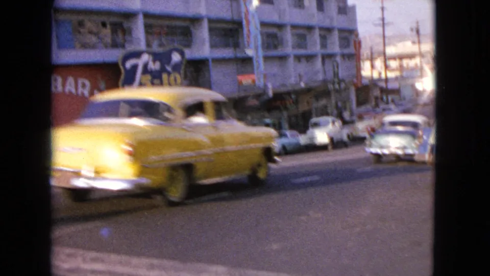 Yellow mid-century car moving through a storefront-lined street in 1963 Barrington, Illinois footage, with parked cars and signs nearby.