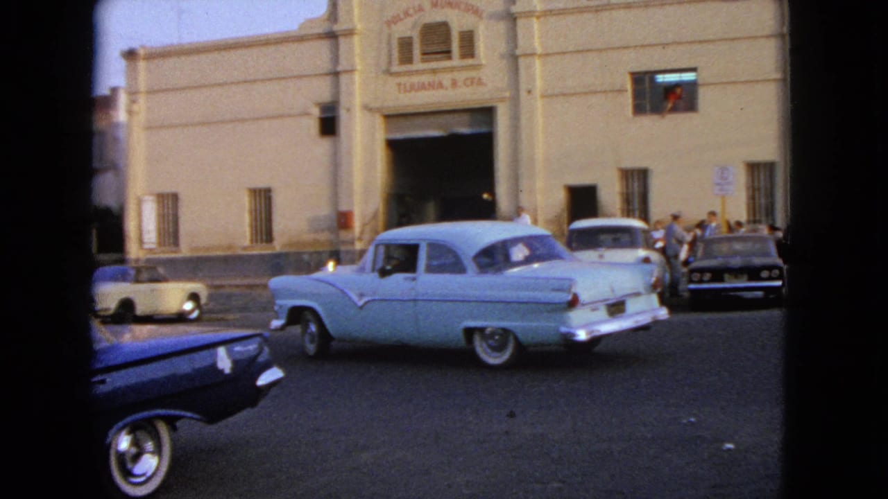 Classic cars pass a beige municipal building with pedestrians near the entrance.