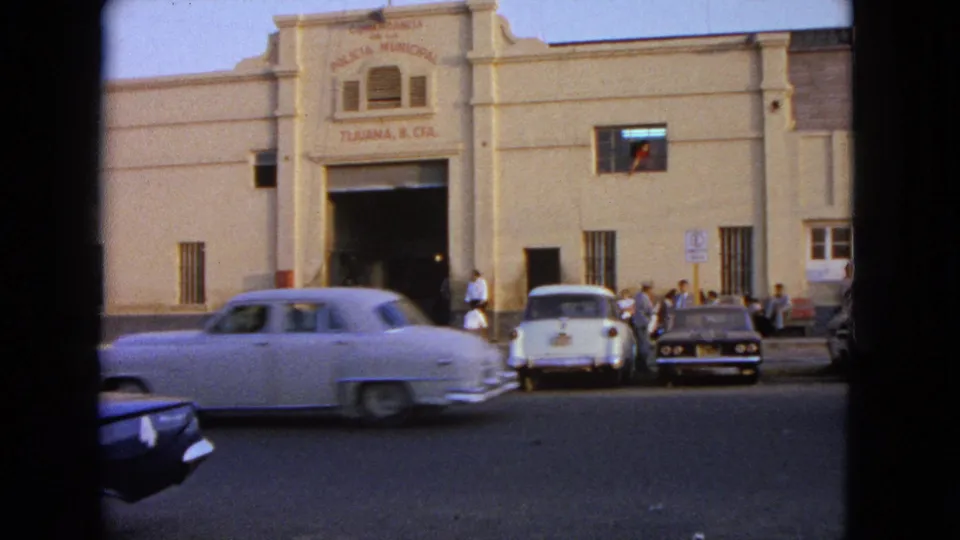 A light-colored classic sedan blurs across the foreground outside the municipal building, with people and parked cars visible behind it.