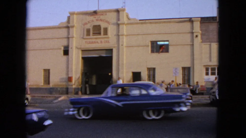 A dark blue 1950s-style car drives past a beige municipal building labeled Policia Municipal, with pedestrians gathered at the entrance.