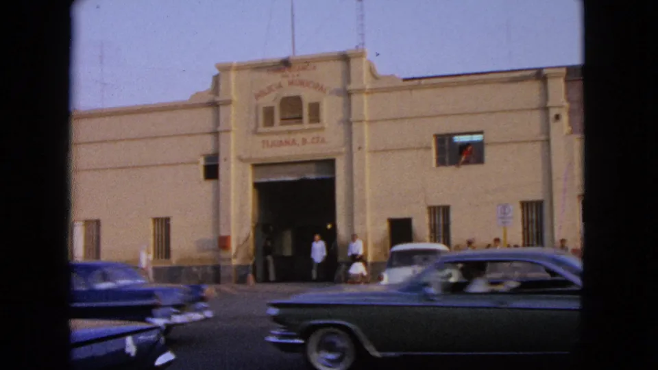 1960s 8mm street view with classic cars passing a beige building signed Policia Municipal and Tijuana, B. C., Mx.