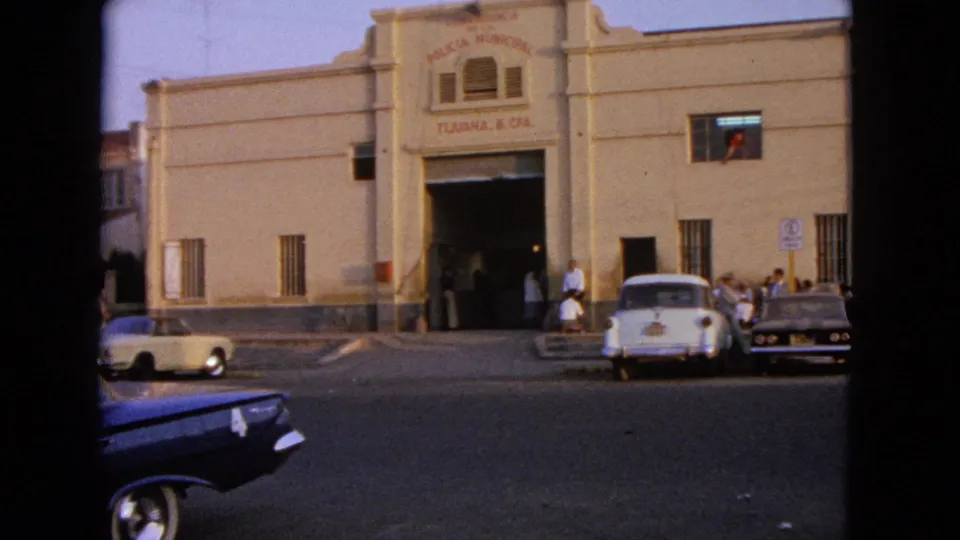 1960s 8mm frame showing a signed municipal facade, parked classic cars, pedestrians, and moving traffic in the street.