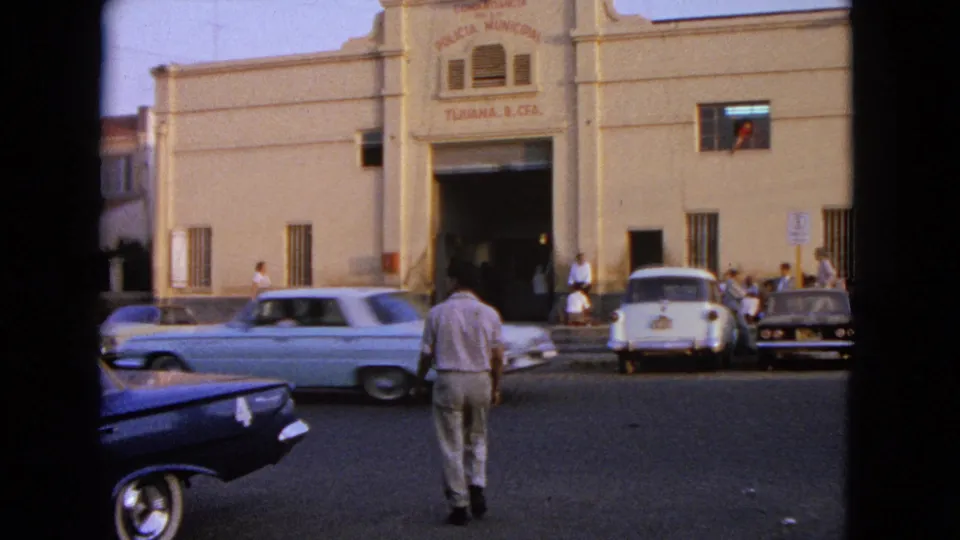1960s 8mm street scene with a man crossing, a light-colored car passing, parked cars, and a signed municipal facade behind him.