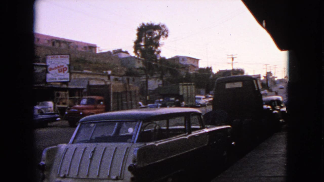 1963 Barrington sidewalk view with a parked station wagon, dark vehicle, distant cars, utility poles, and commercial buildings.