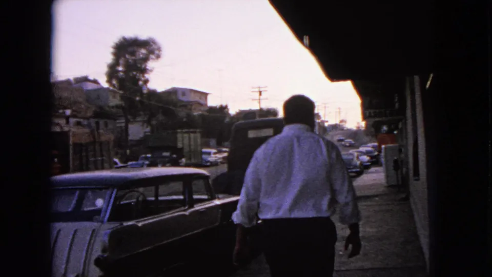 A man in a white shirt walks away from the camera on a Barrington sidewalk beside parked 1960s cars.