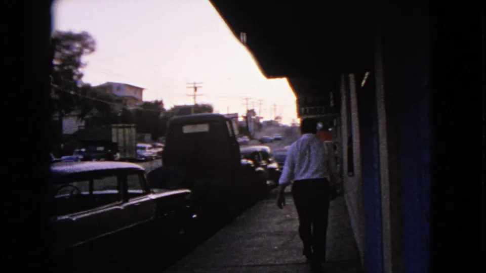The man continues down the Barrington sidewalk, moving away past a dark vehicle and parked classic cars in 1963 home-movie footage.