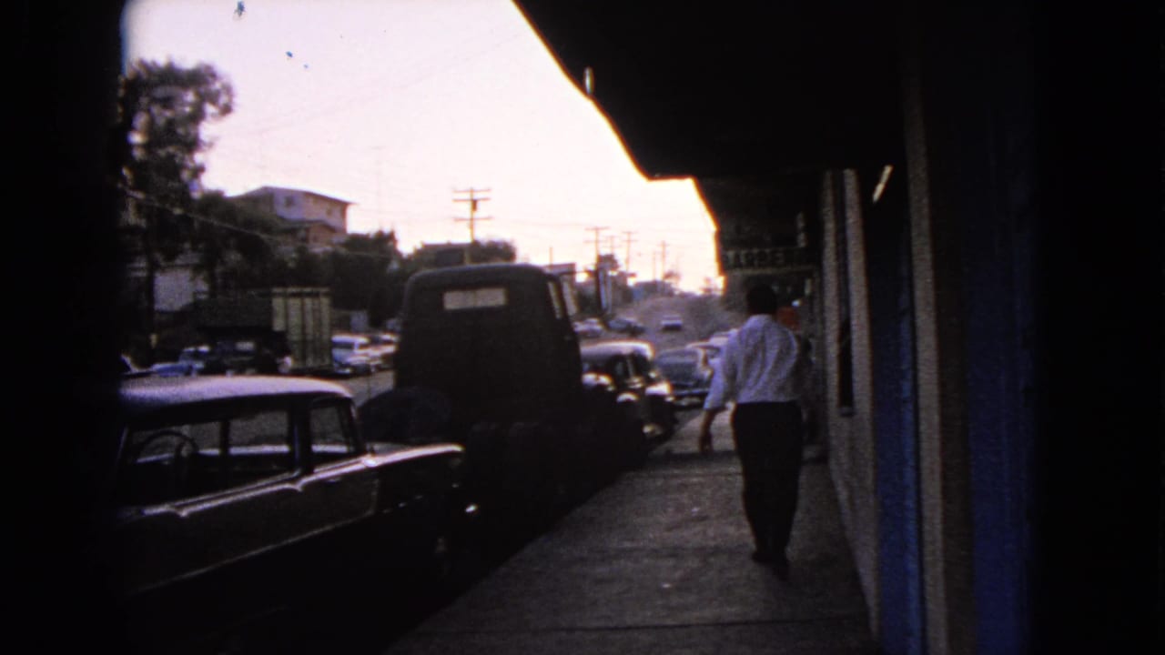 1963 Barrington, Illinois street view with a man walking along a sidewalk past parked mid-century cars and shaded storefronts.
