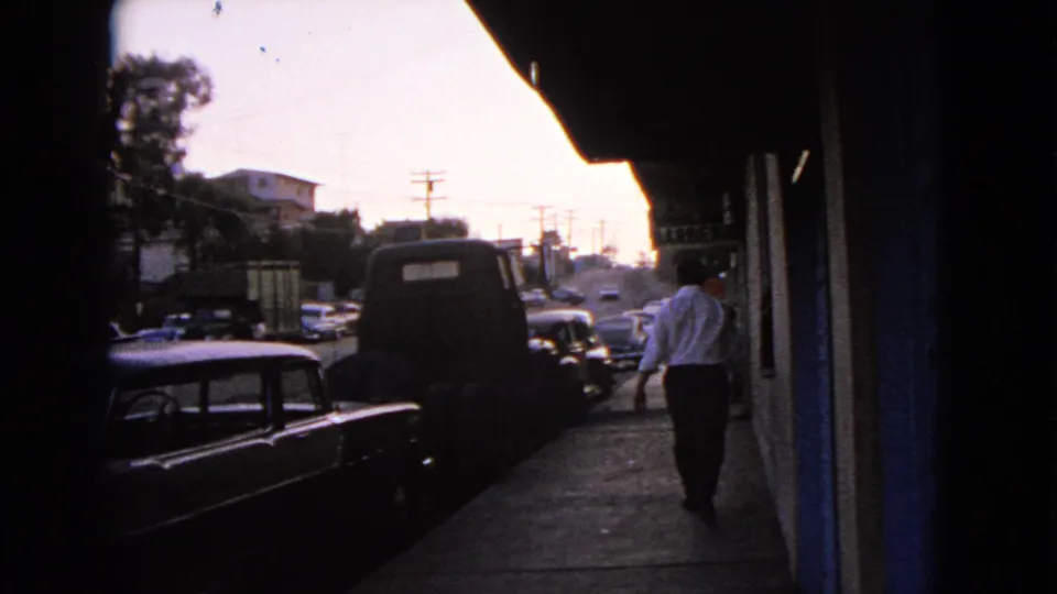 1963 Barrington, Illinois street view with a man walking along a sidewalk past parked mid-century cars and shaded storefronts.