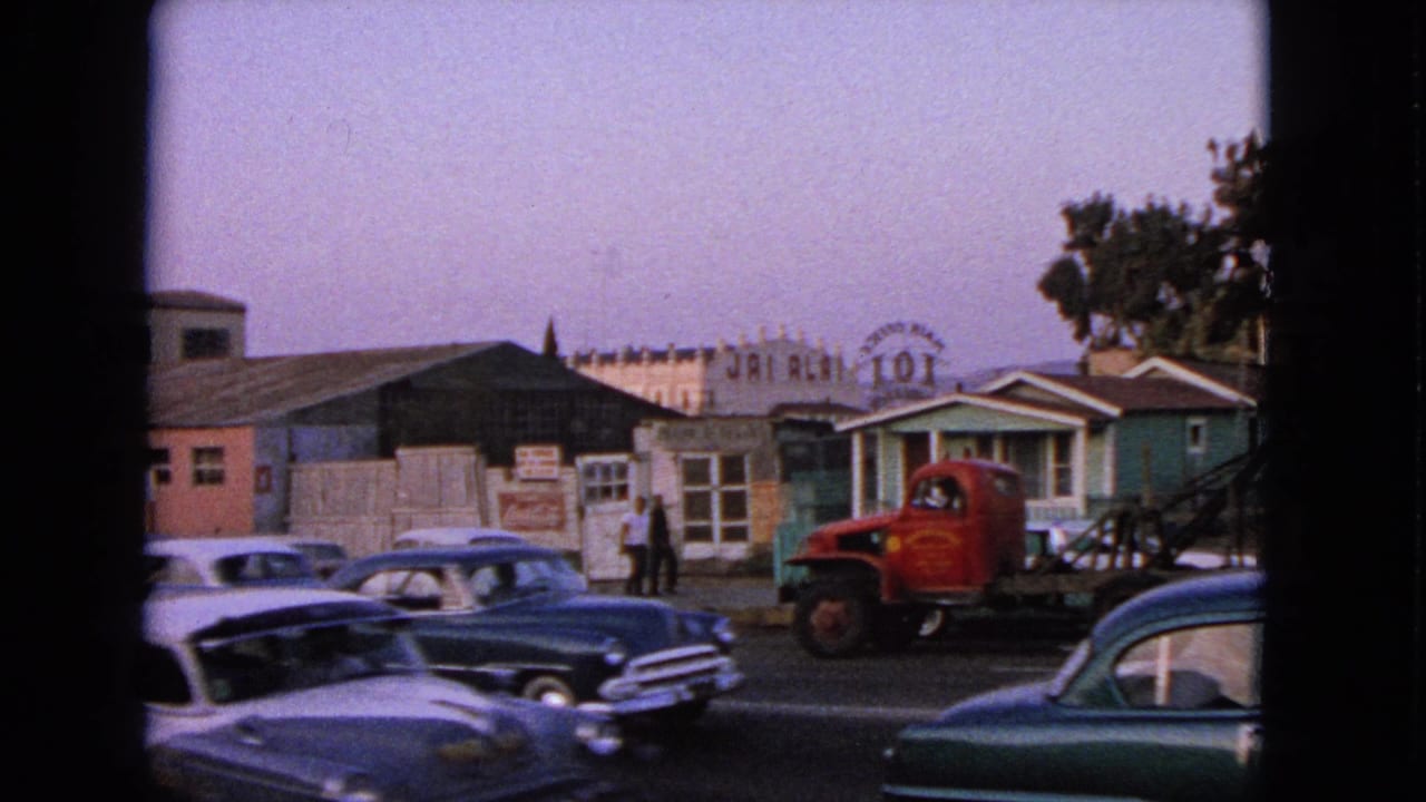 Classic cars pass low buildings near a red tow truck and distant signage.