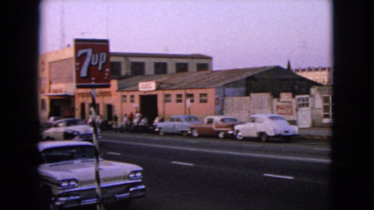 Street scene with a 7-Up sign, parked and passing cars, and industrial buildings across the road.