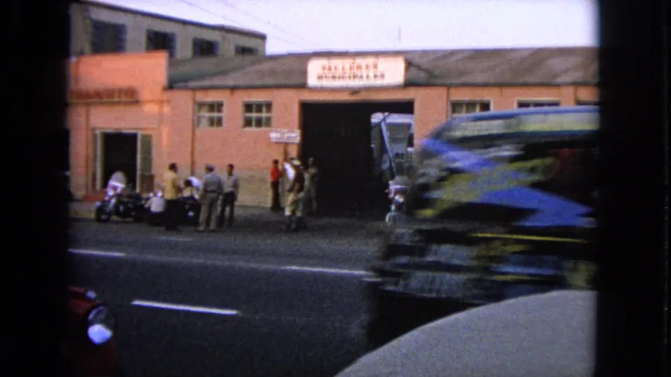 1963 8mm frame showing a truck passing a pink building marked “TALLERES MUNICIPALES,” with pedestrians and motorcycles outside.