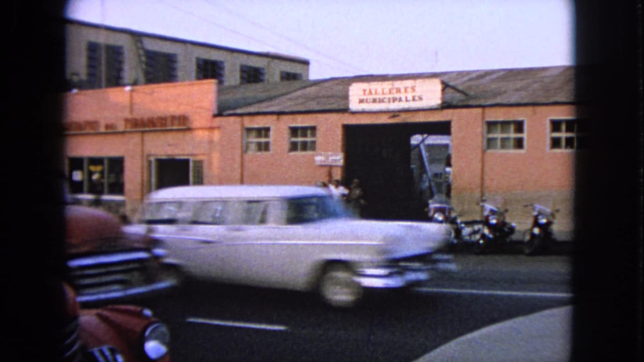 Classic cars pass a municipal workshop building with pedestrians and motorcycles nearby.