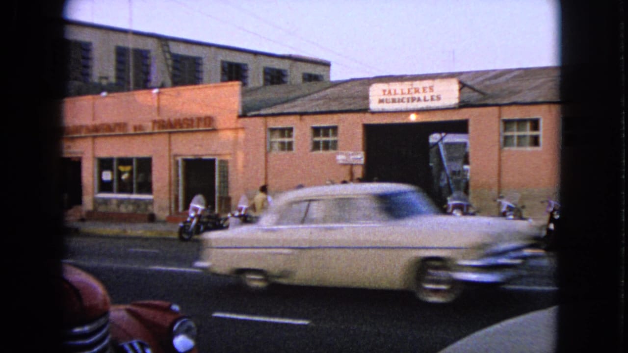 Light-colored sedan passes in front of workshops with motorcycles and pedestrians by the curb.