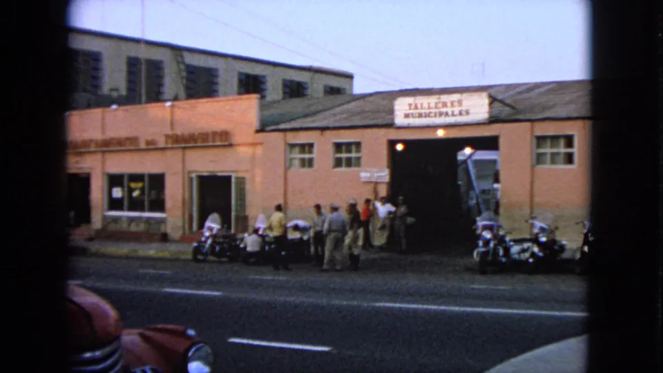 Pedestrians stand outside a municipal workshop in the Barrington clip, with motorcycles parked near the curb and traffic framing the scene.