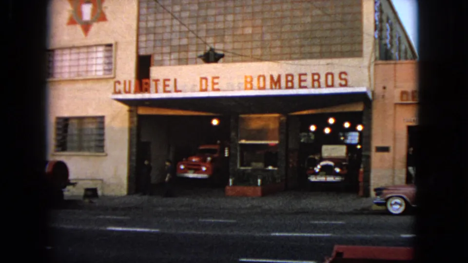 The clip cuts to a fire station marked in Spanish, with red fire vehicles parked inside open garage bays.