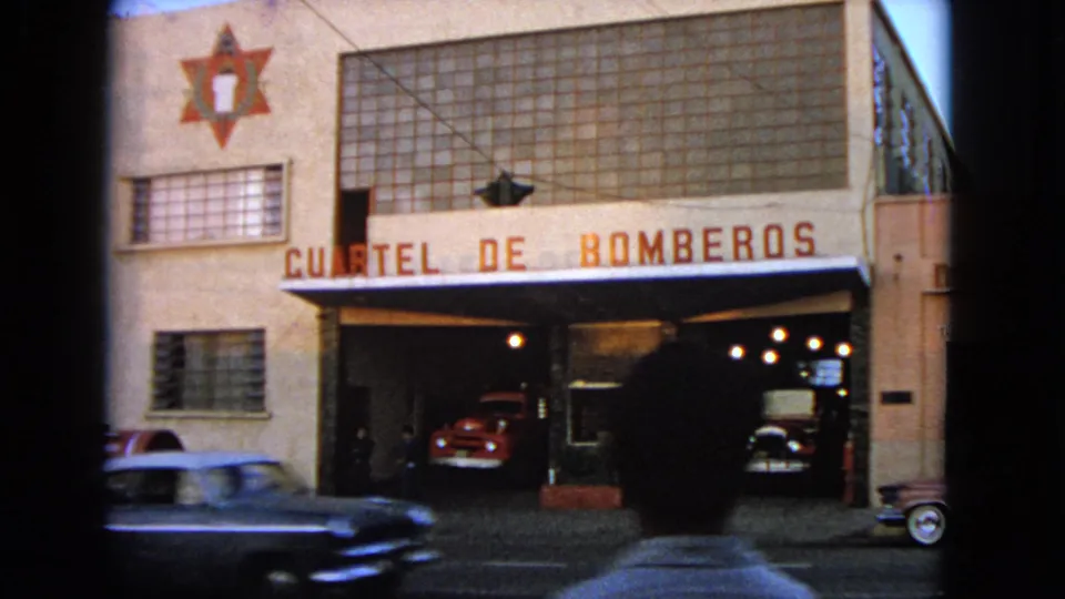 1963 home-movie frame identified as Barrington, Illinois, with a passing car and foreground silhouette in front of the fire station and truck bays.