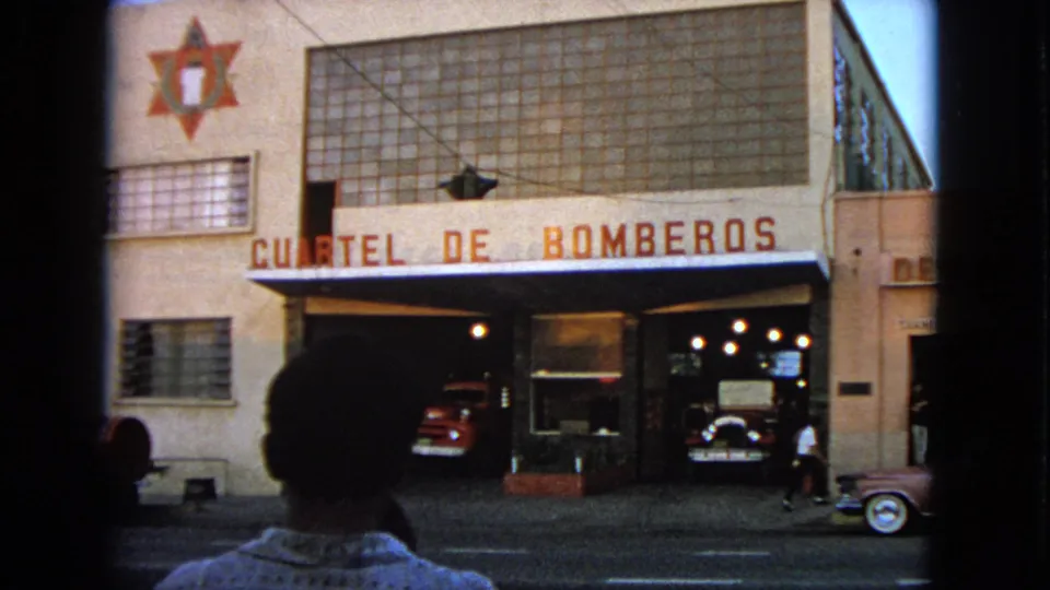 End frame from 1963 Barrington home-movie footage showing a foreground figure, fire station bays, red trucks, and a pink car near the curb.