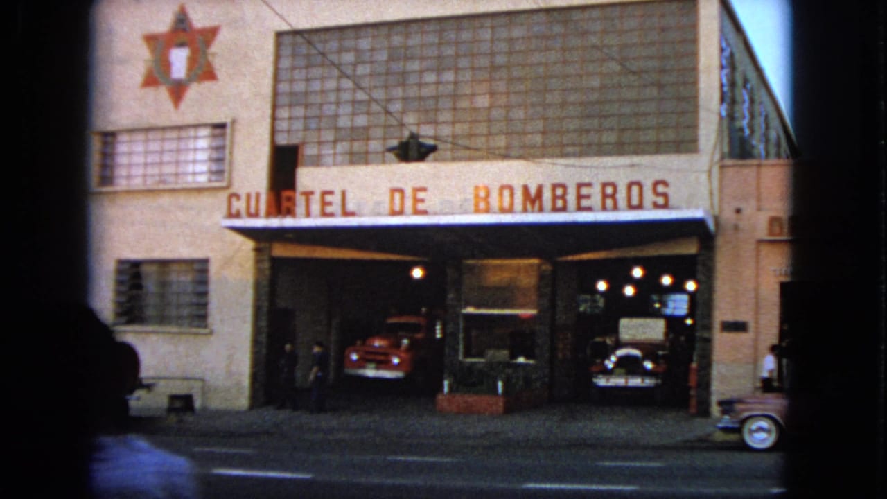 1963 8mm frame showing a fire-station facade with Spanish text, a star emblem, people near the entrance, and classic cars in the bays.