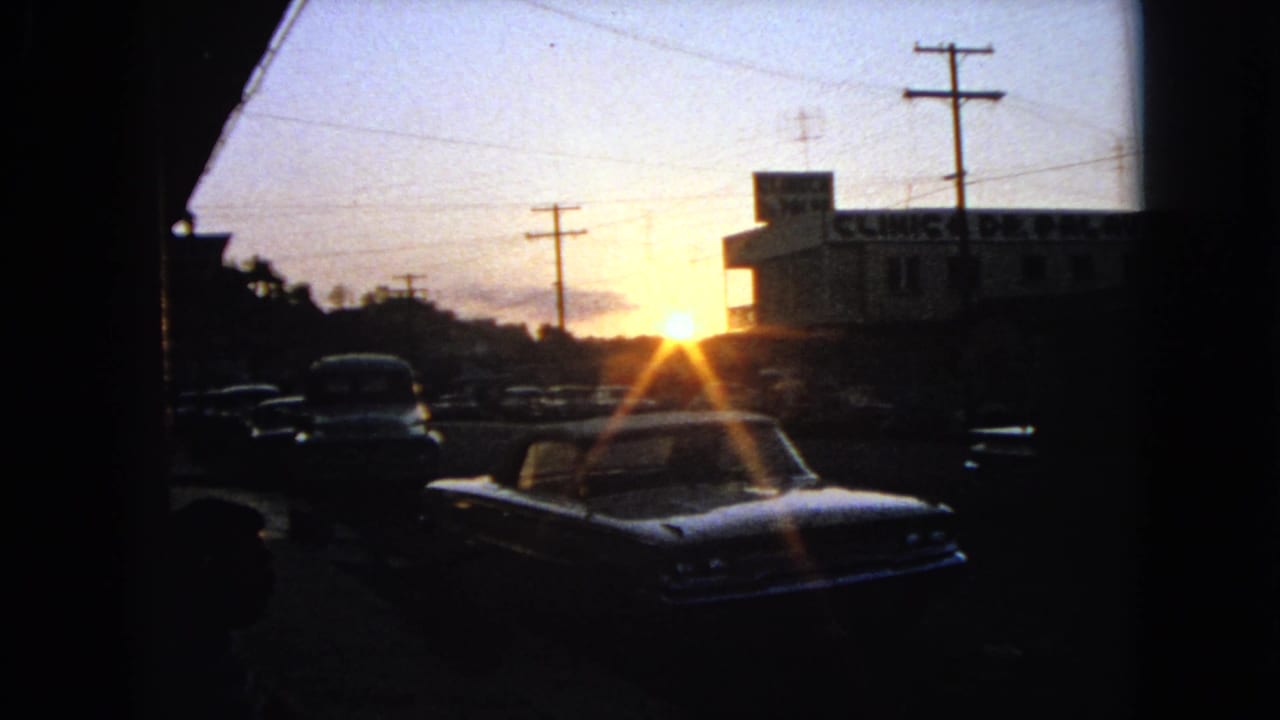 1963 8mm street view at sunset with classic cars in shadow, utility poles, and a building sign partially visible on the right.