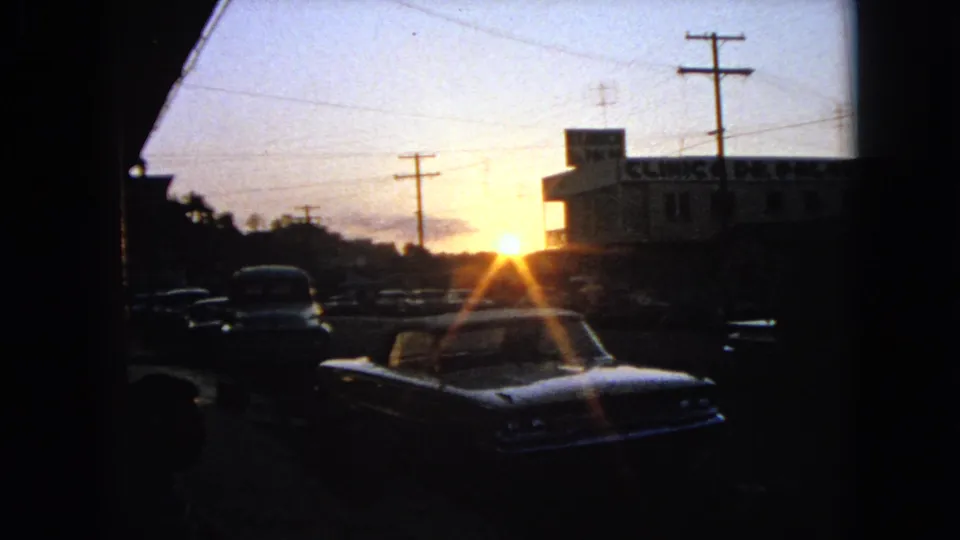 1963 8mm street view at sunset with classic cars in shadow, utility poles, and a building sign partially visible on the right.