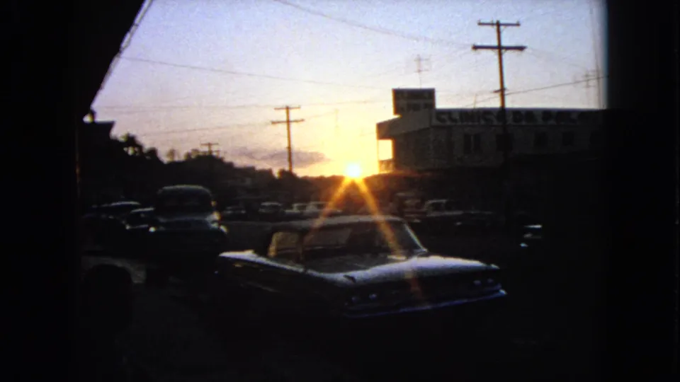 1963 8mm frame of a sunset street with a classic car foreground, utility poles, and a commercial building edge in silhouette.